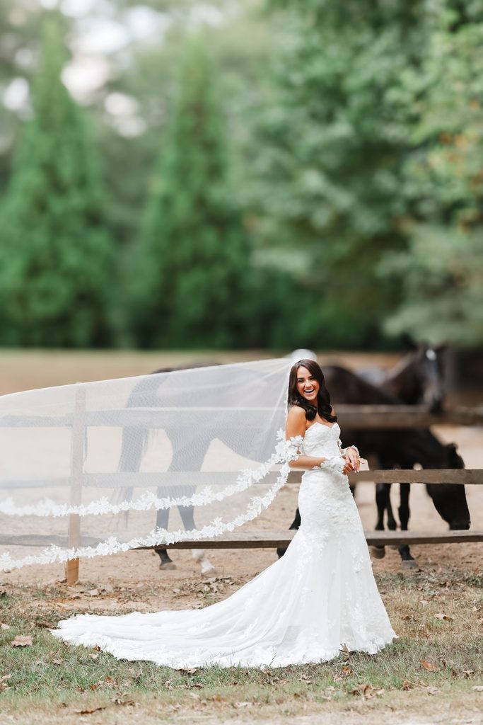 A bride in a white lace wedding gown stands in front of a wooden fence with horses, her long veil blowing in the wind.