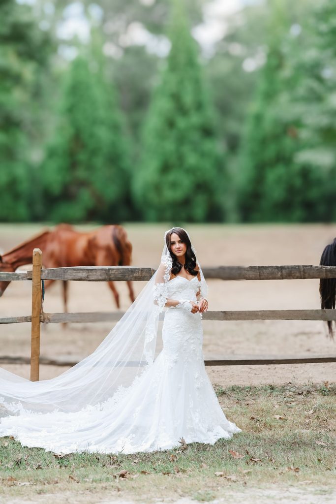 A bride in a lace mermaid wedding dress and veil stands in front of a wooden fence with a horse in the background.