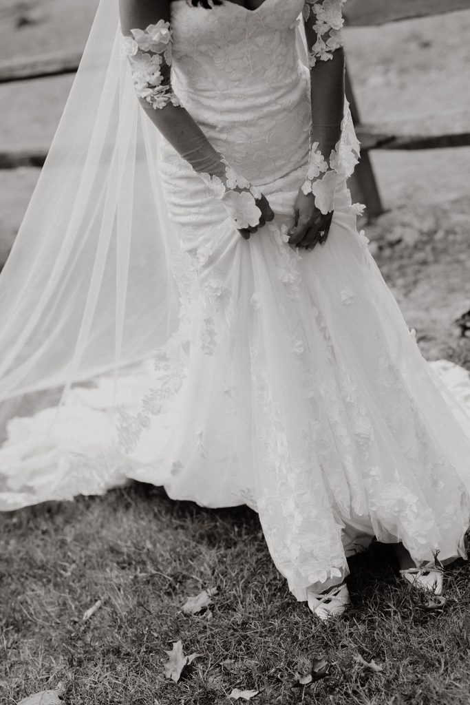 A bride in a floral-appliqué lace gown with long sleeves holds the front of her skirt, with a long veil trailing behind her on the grass.