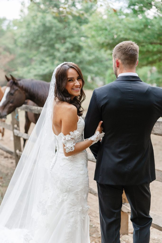 A smiling bride in a white wedding gown and veil looks back at the camera while standing with her groom near a wooden fence with horses in the background.