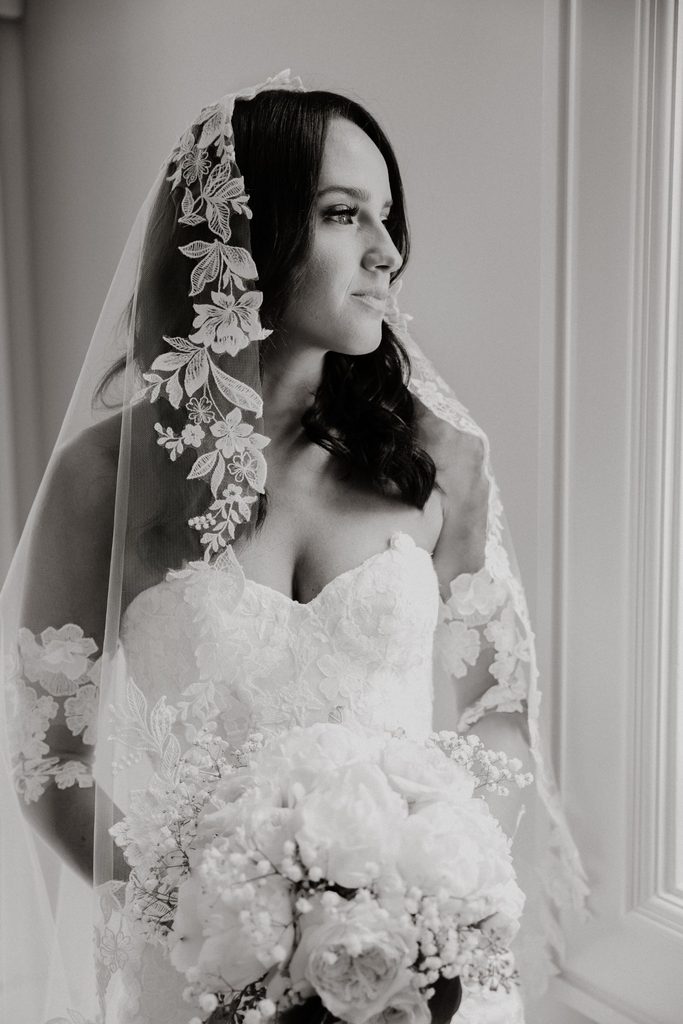A bride in a strapless gown and lace-trimmed veil looking out a window while holding a bouquet of white flowers.