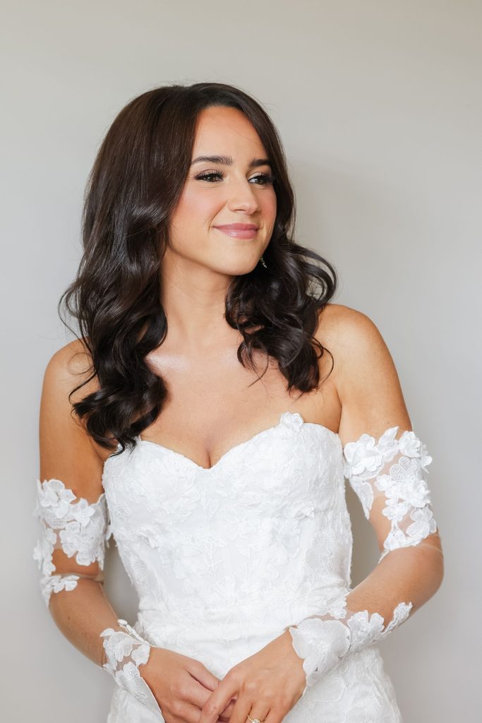 A bride in a strapless lace wedding gown with matching detachable sleeves, looking away from the camera.