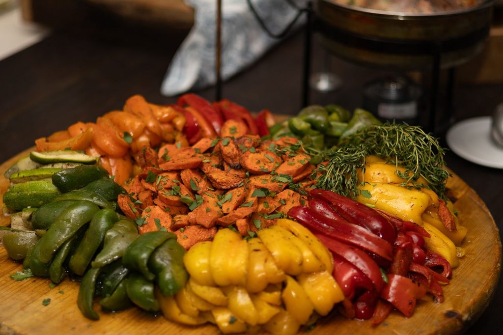 A wooden platter piled with roasted sliced carrots surrounded by strips of green, yellow, orange, and red bell peppers, garnished with fresh herbs.