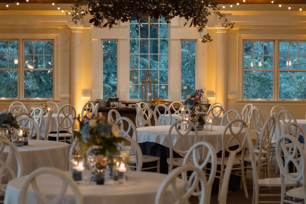 A banquet hall with round tables covered in white tablecloths, surrounded by white chairs, and a buffet table set up in front of a large window with greenery hanging above.
