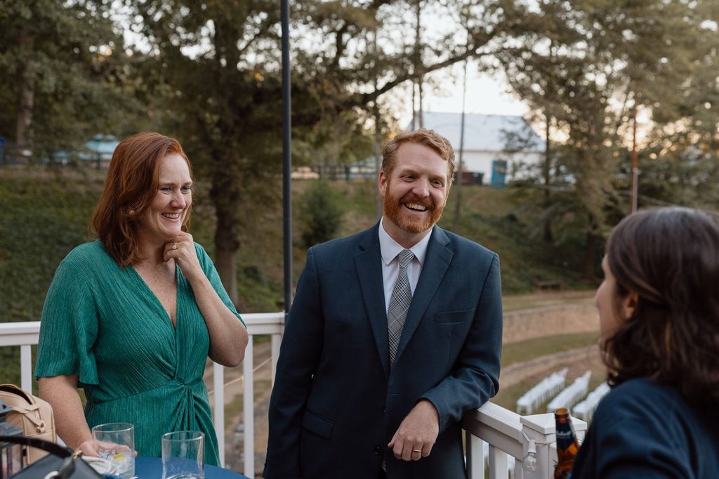 A smiling man in a suit and a woman in a green dress talk to someone off-camera on a deck.