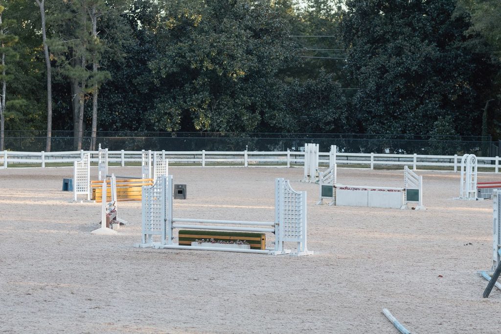 An empty horse jumping arena with various white, yellow, and green obstacles set up on the sand.