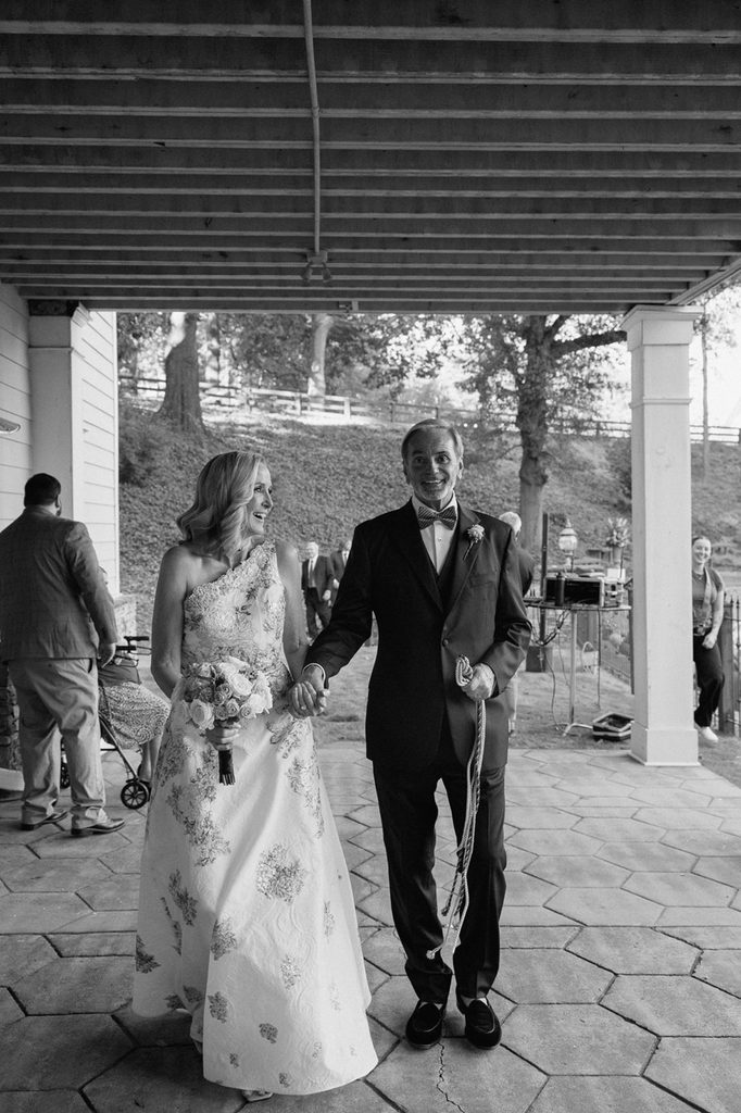 A bride in a patterned gown and a groom in a tuxedo walk hand-in-hand together on a patio.