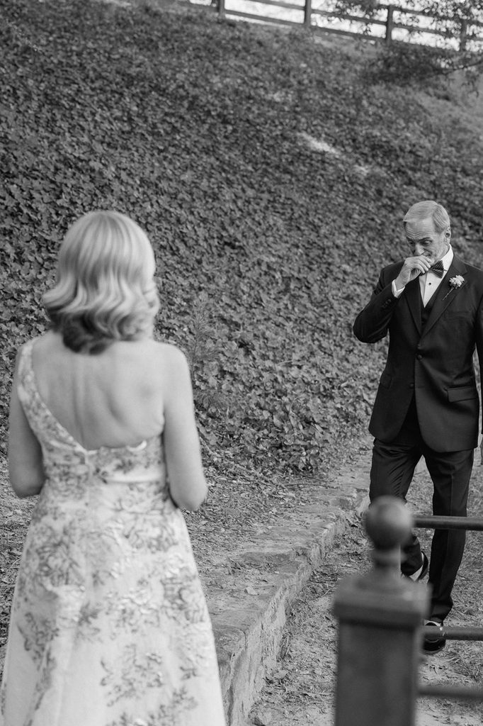 A man in a tuxedo wipes a tear from his eye as he looks toward a woman in a floral dress.