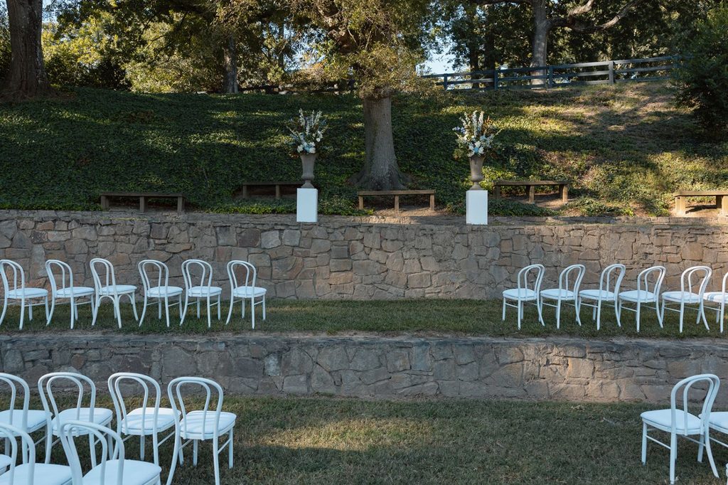 Rows of white chairs arranged for an outdoor wedding ceremony in front of a stone wall with two floral pedestals.