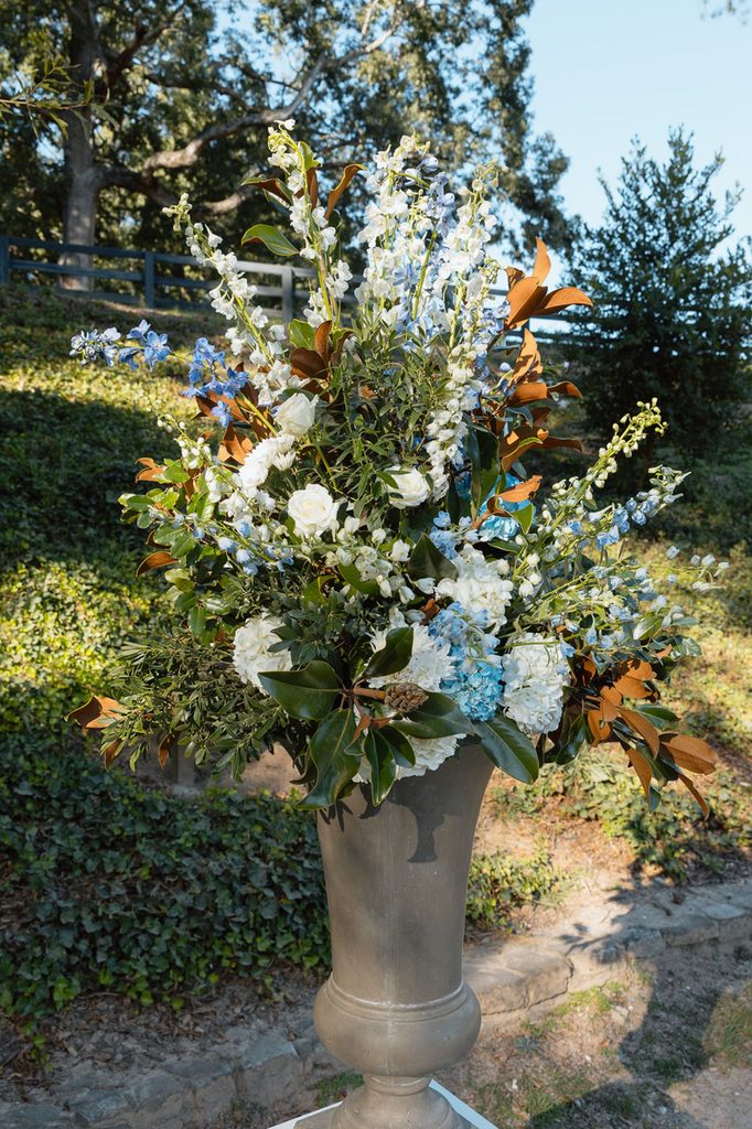 A tall, flared vase filled with an arrangement of white roses, blue delphiniums, white hydrangeas, and magnolia leaves.