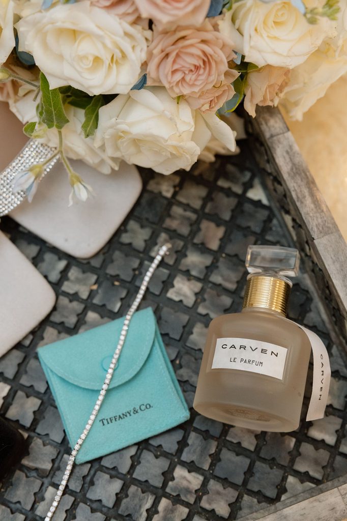 Carven Le Parfum perfume bottle, a Tiffany & Co. jewelry pouch, a thin diamond bracelet, and a bouquet of white and blush roses on a decorative tray.