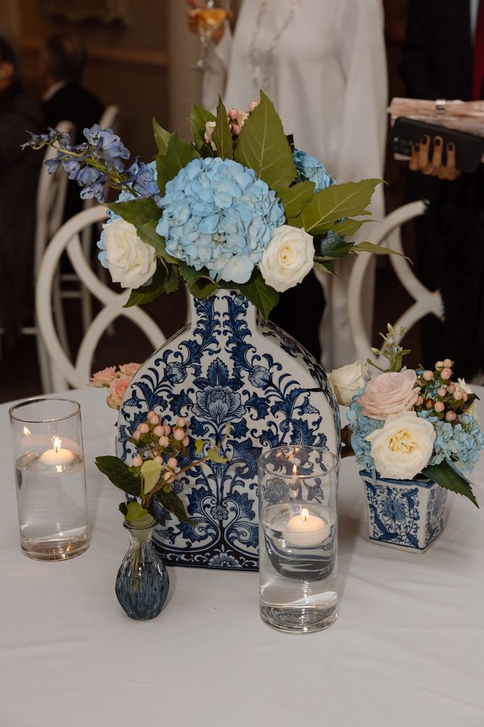 A table centerpiece featuring blue and white patterned vases filled with blue hydrangeas, white roses, and greenery, surrounded by two glass cylinders containing floating candles.