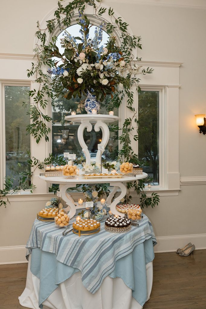 A three-tiered dessert display stand covered in assorted pastries and cupcakes, set on a blue-striped tablecloth in front of a large arched window adorned with greenery.