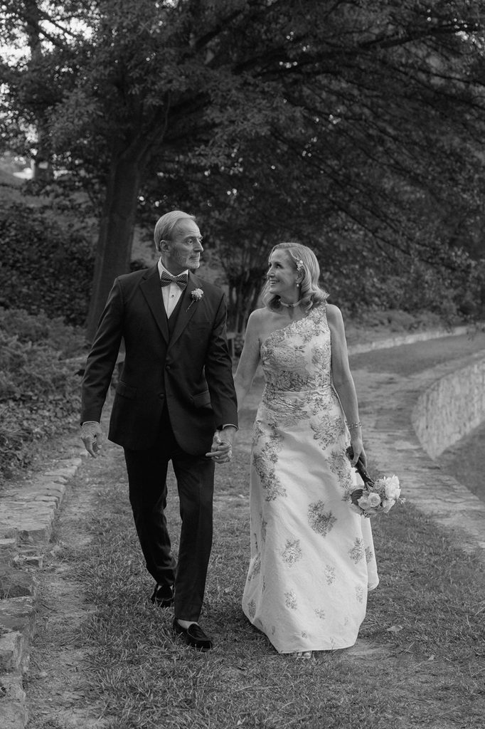 A man in a suit and a woman in a floral gown hold hands while walking along a garden path.