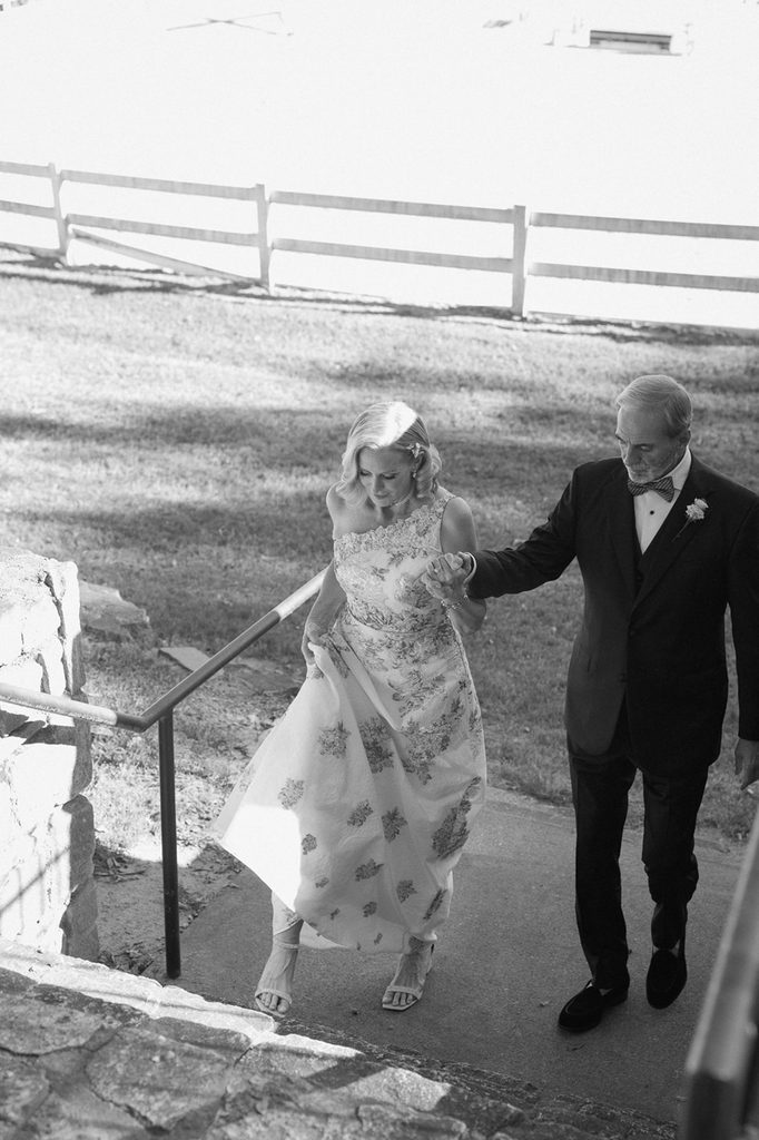 A man in a tuxedo holding hands with a woman in a floral one-shoulder gown as they walk up concrete stairs together.