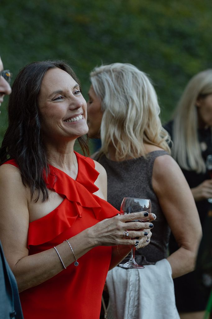 A smiling woman in a red ruffled dress holds a glass of red wine while talking to others at an outdoor gathering.
